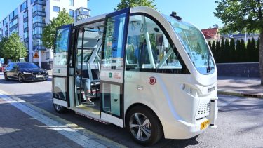 Fehér önvezető transzferbusz áll meg városi környezetben, nyitott ajtóval, utasforgalmi tesztüzem közben.